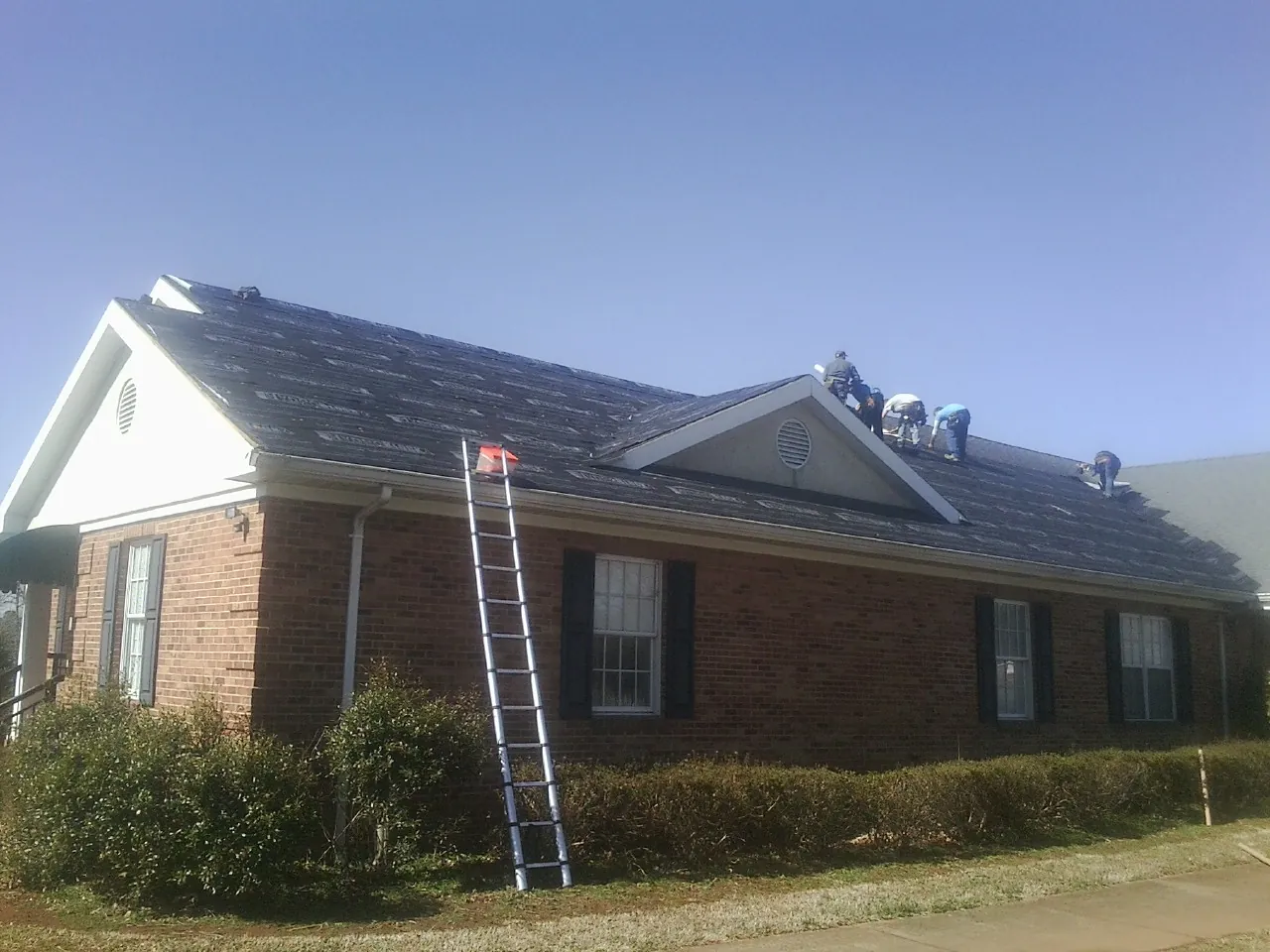 Crew tearing shingles off a brick ranch roof with a ladder set — Metal Roof Repair work by our crew in North Sutton