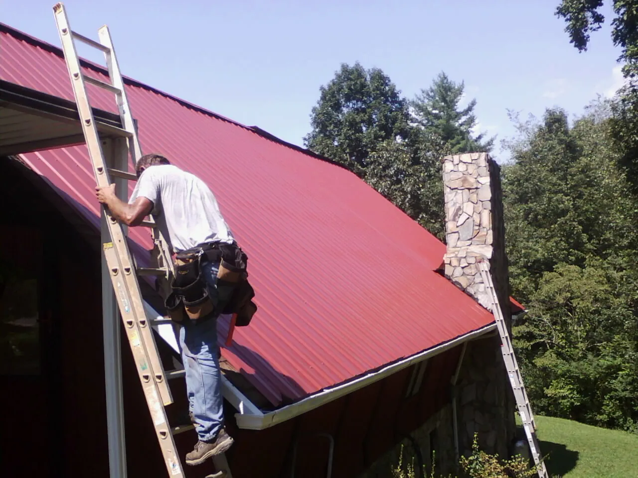 Roofer on a ladder beside a red metal roof and stone chimney — Soffit & Fascia Repair by Roofing Brothers in North Sutton
